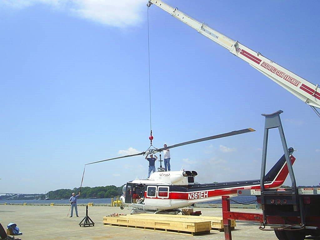 A helicopter suspended from a mobile crane being lowered onto timber dunnage at a dock.
