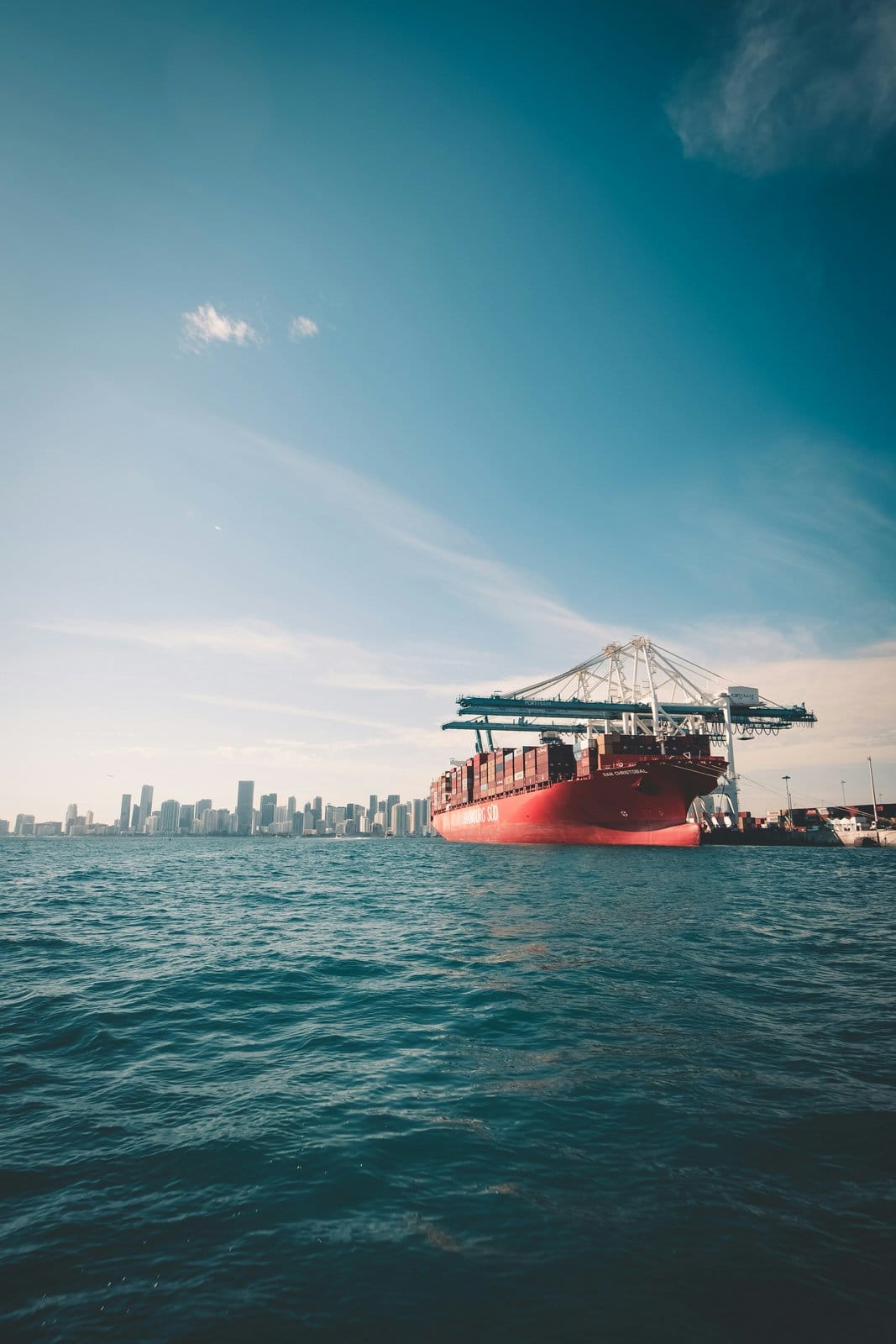 Container ship alongside gantry cranes with the Miami skyline behind it, representing the ocean leg of the Miami to Brazil trade lane