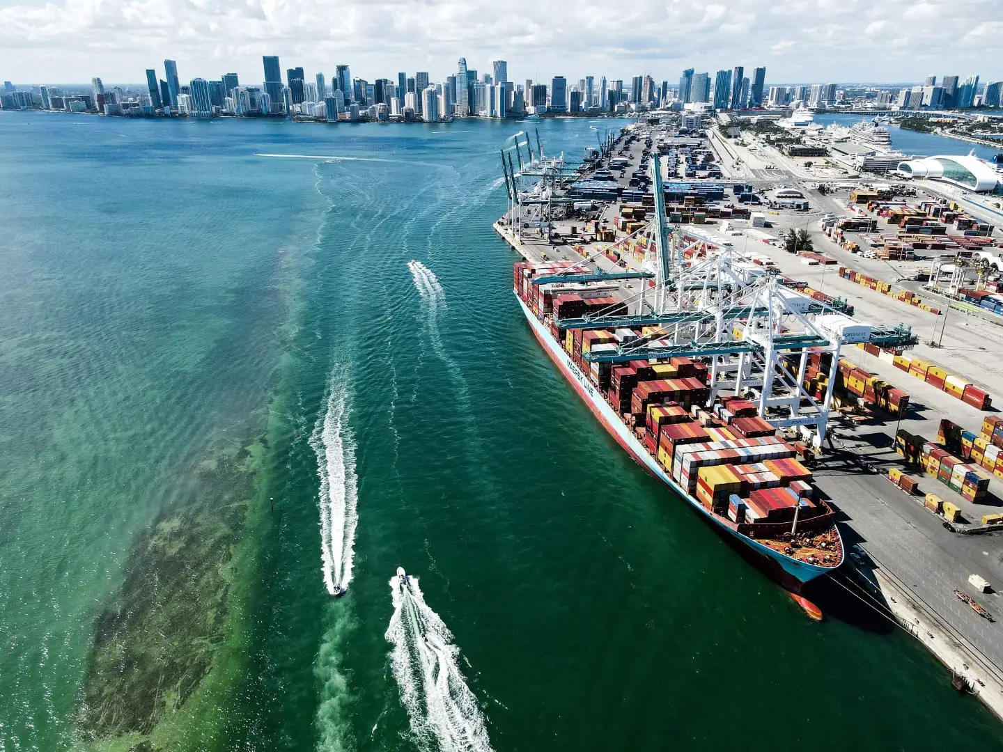 Vista aérea de un portacontenedores atracado en PortMiami, con grúas pórtico, el canal de navegación y los edificios de Miami al fondo