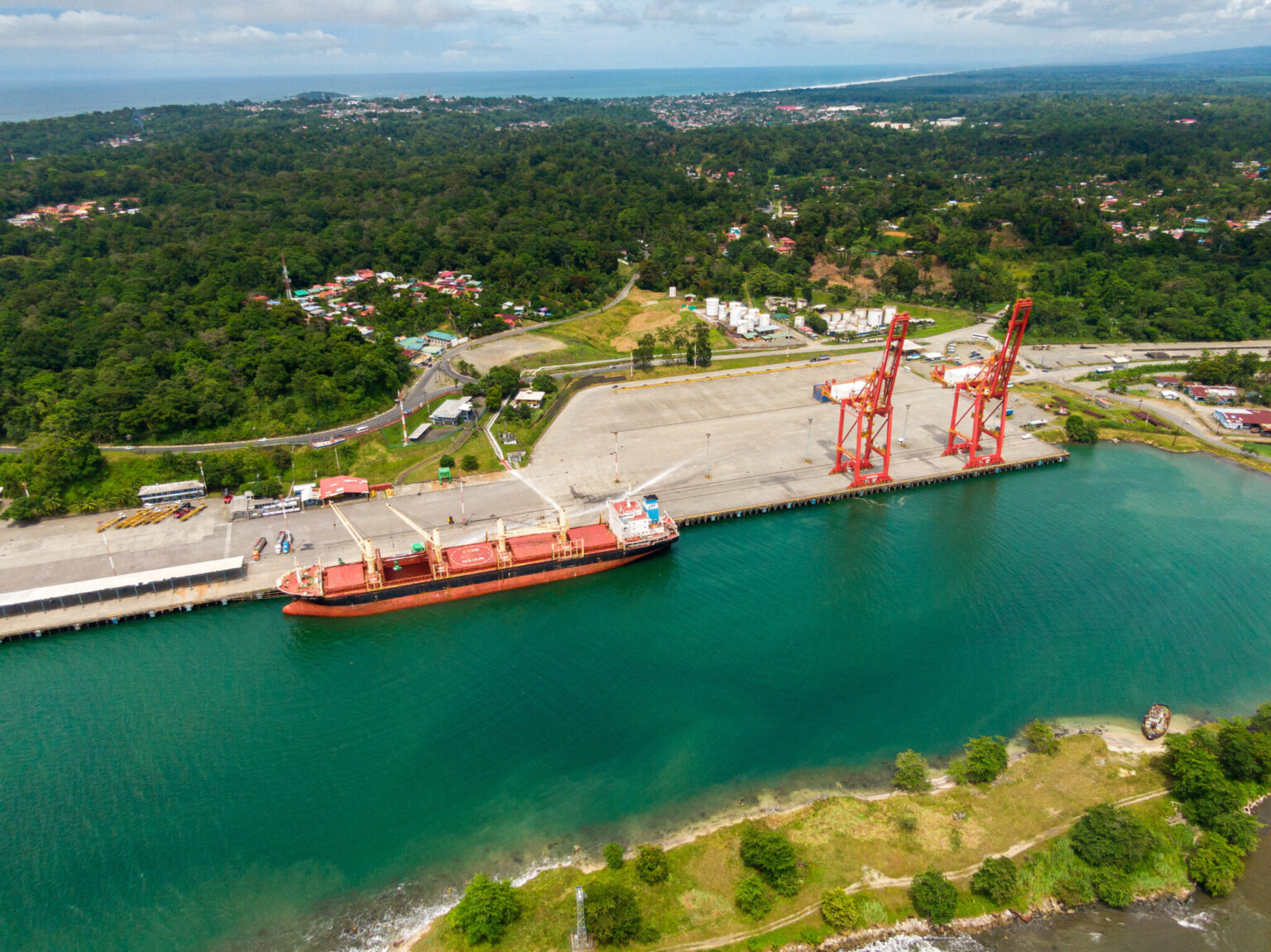 Aerial view of the port of Moín in Limón, Costa Rica, with a cargo ship at the berth, red cranes on the pier and rainforest behind
